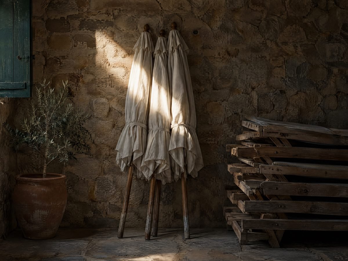 Three closed white parasols and a stack of wooden sun loungers stored in a stone room of a hotel in Rhodes in winter, lit by a single beam of sunlight from a window with a green shutter