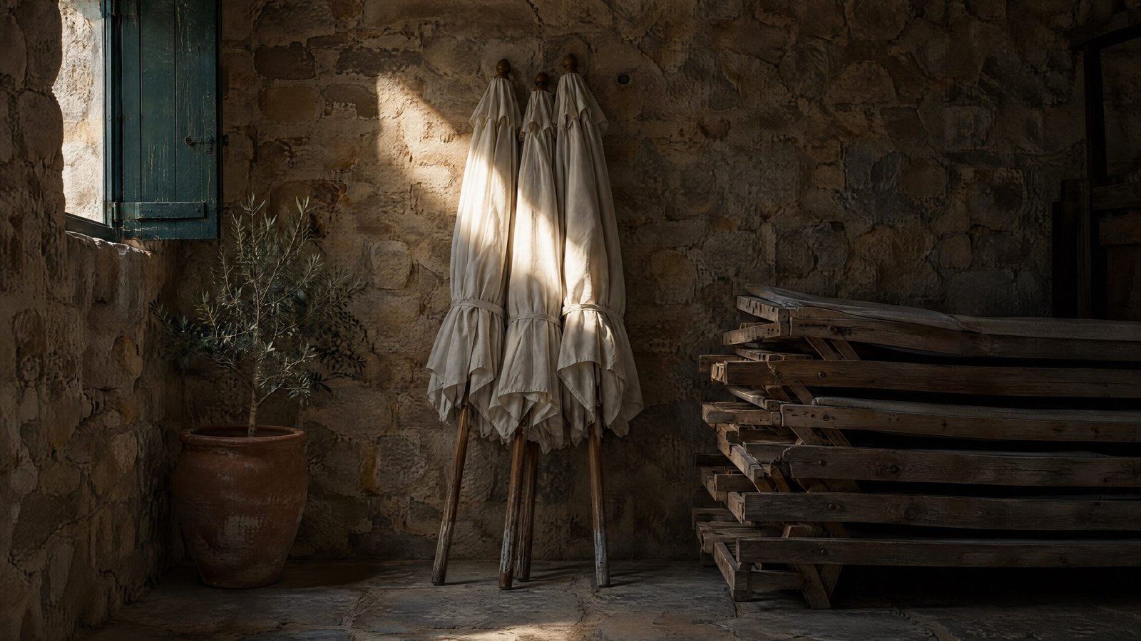 Three closed white parasols and a stack of wooden sun loungers stored in a stone room of a hotel in Rhodes in winter, lit by a single beam of sunlight from a window with a green shutter